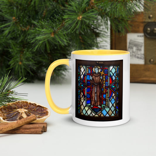 A ceramic mug with a stained glass design featuring a religious figure, placed on a table with a pine tree in the background and some cinnamon sticks to the side.